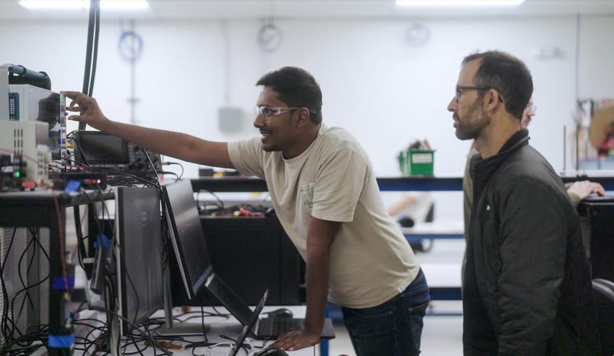 Two men in foreground review a device showing waveforms; a man in the background is mostly hidden. Two men in foreground review a device showing waveforms; a man in the background is mostly hidden.