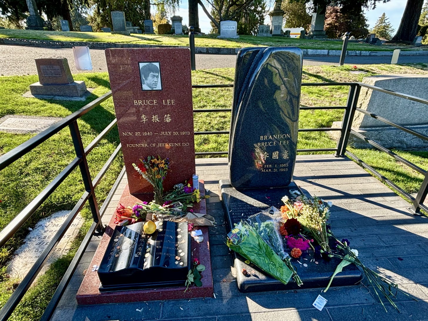 color photo of two grave markers for Bruce Lee and Brandon Lee, with flowers, coins, and other things left as offerings