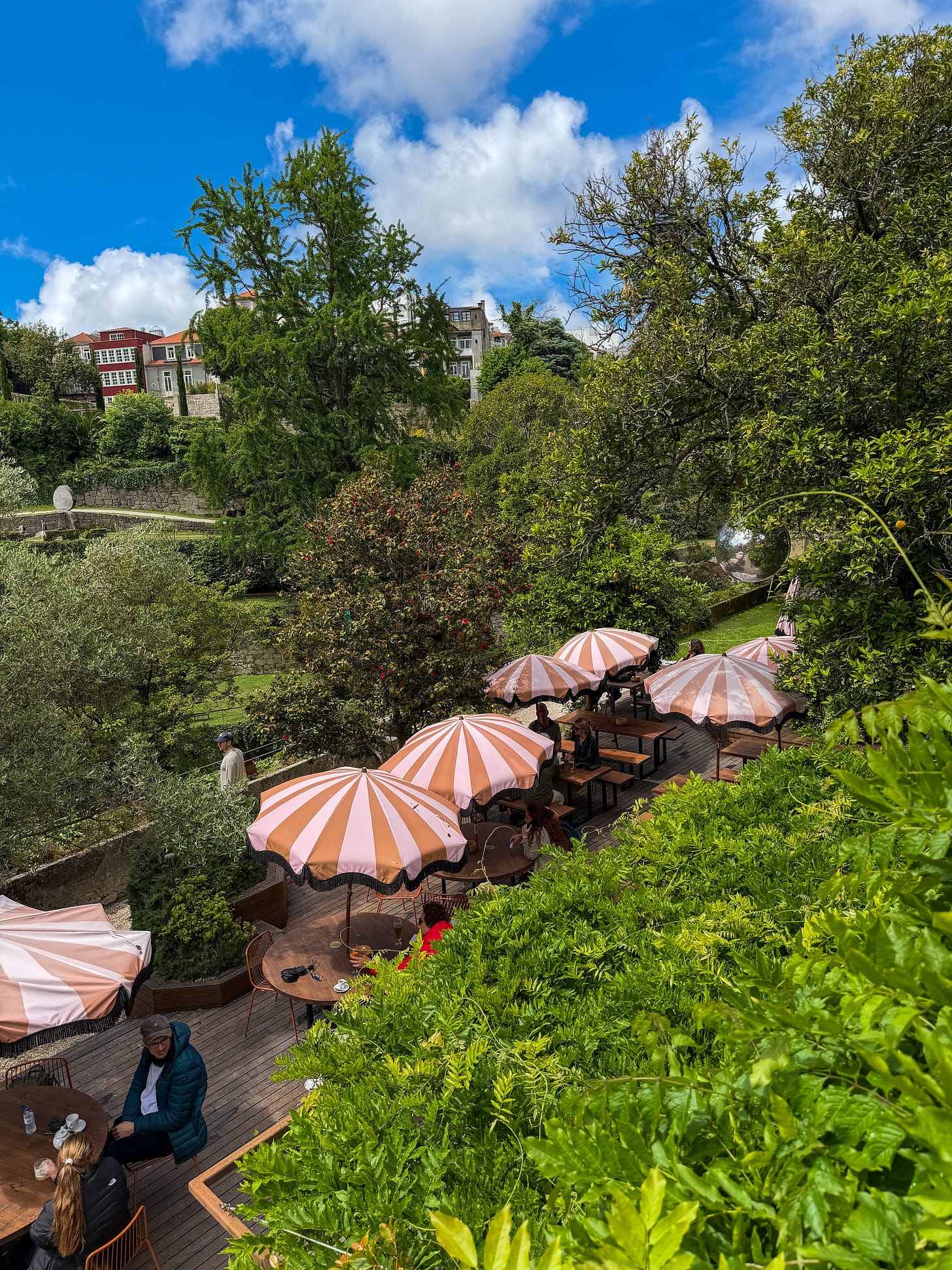 The lush green garden terrace at Bosco bar in Porto, Portugal, with pink and white striped parasols and wooden tables set into a hillside garden above the Douro River