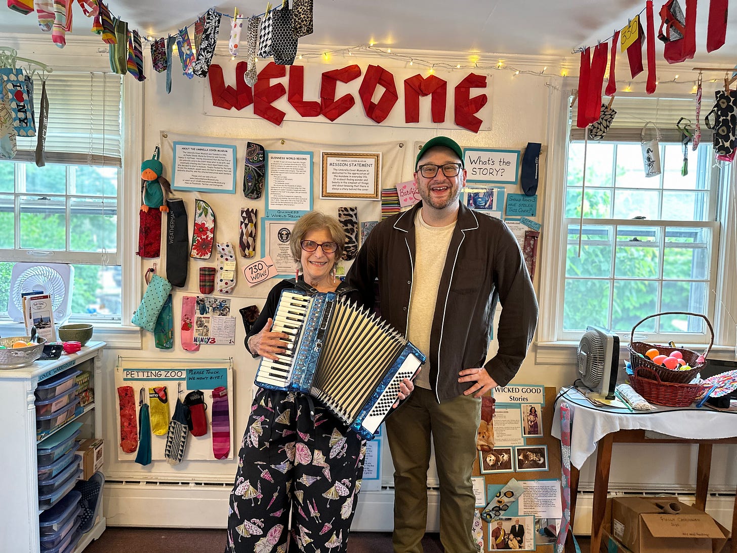 Me and Nancy 3. Hoffman (who is holding an accordion) in the Umbrella Cover Museum. We are standing in front of a WELCOME sign made out of umbrella covers and a few dozen other umbrella covers of various designs.