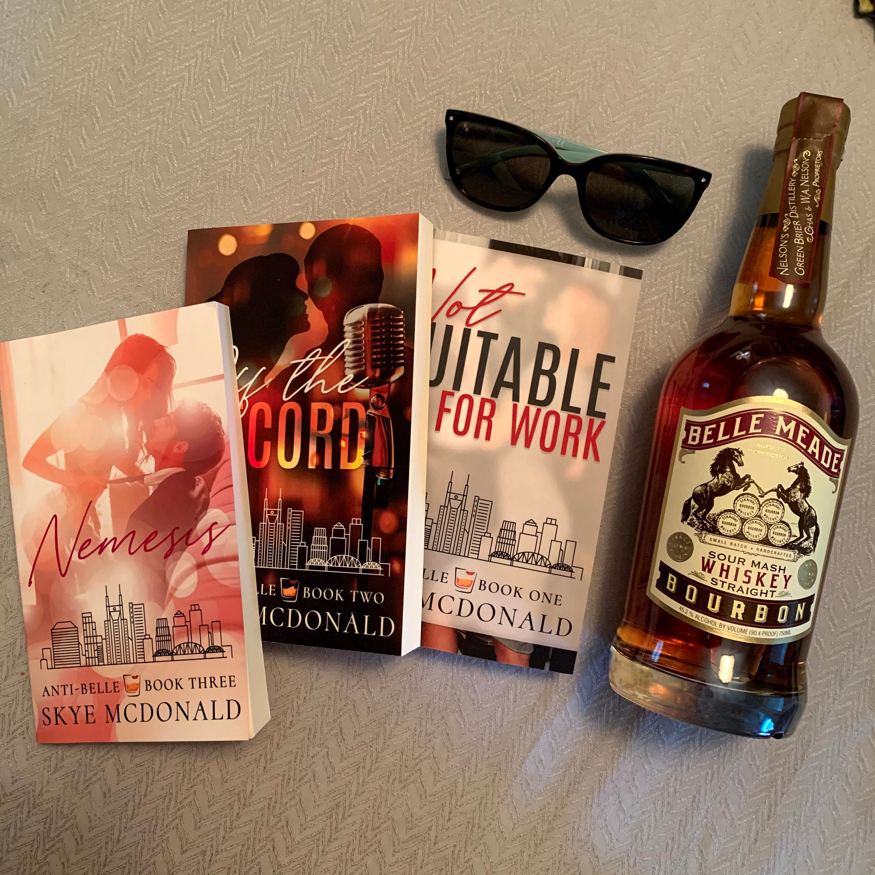 Skye McDonald’s books: Nemesis, Off the Record, and Not Suitable for Work pictured with sunglasses and a bottle of Belle Meade whiskey