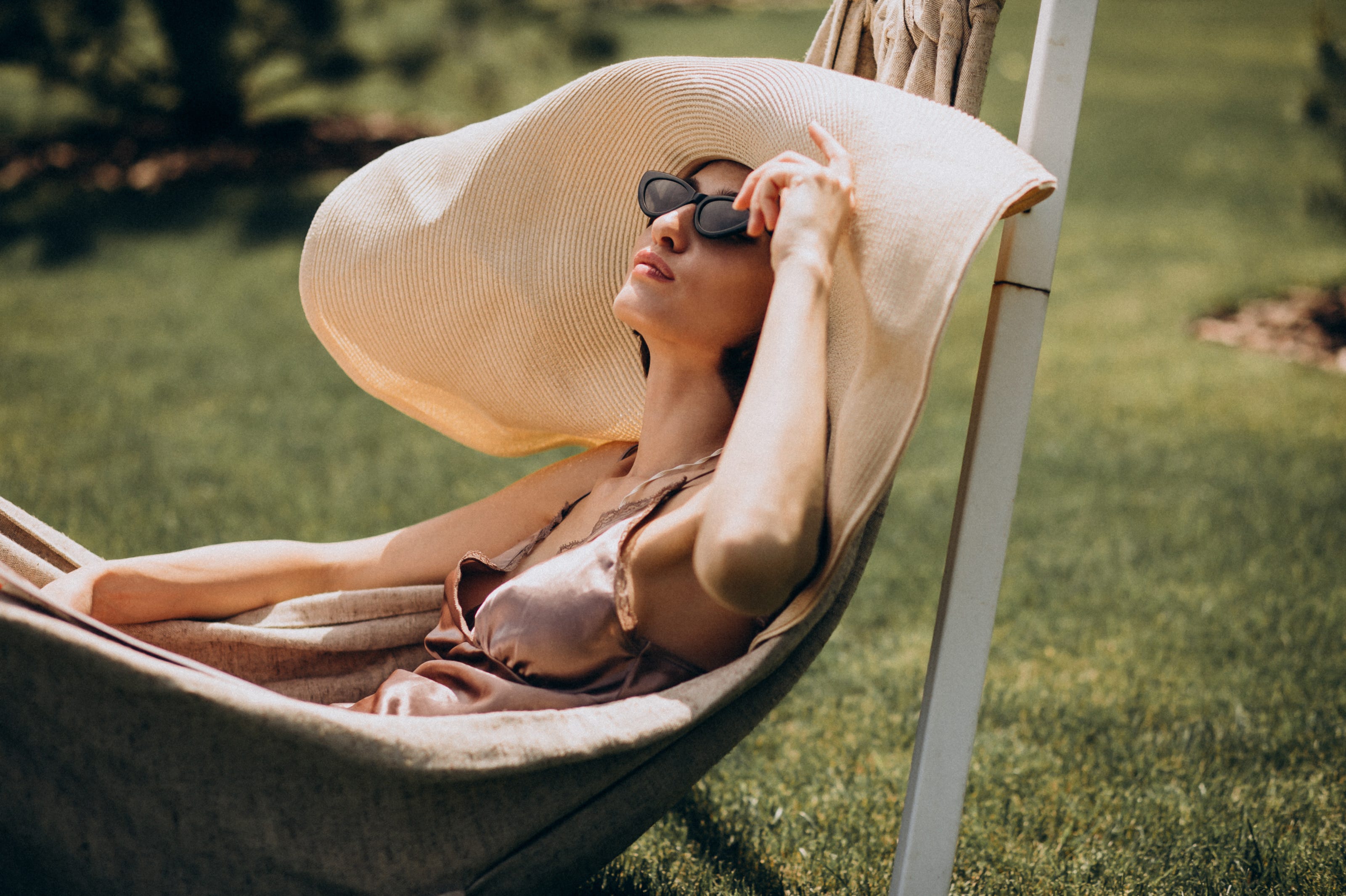 mulher com grande chapéu e óculos de sol a descansar numa rede num jardim, enquanto apanha sol