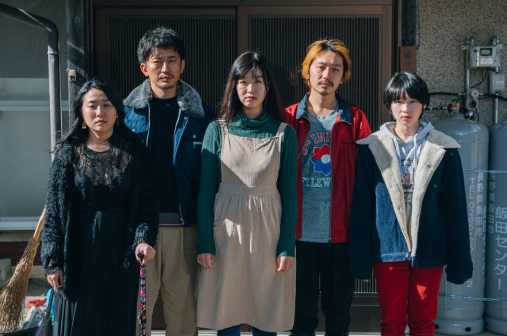 A still from a film showing five people standing together looking at the camera