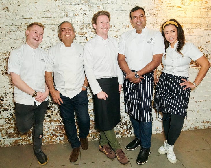 Five chefs lined up against a white brick wall posing for a photo