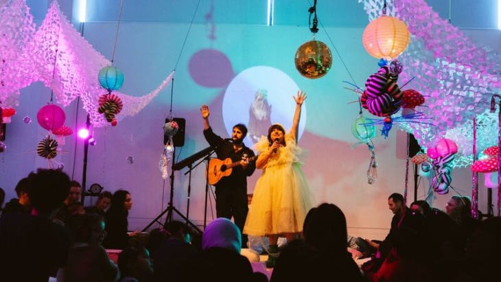 Two performers on stage, one dressed in black with a guitar, the other in a yellow dress, in front of an audience