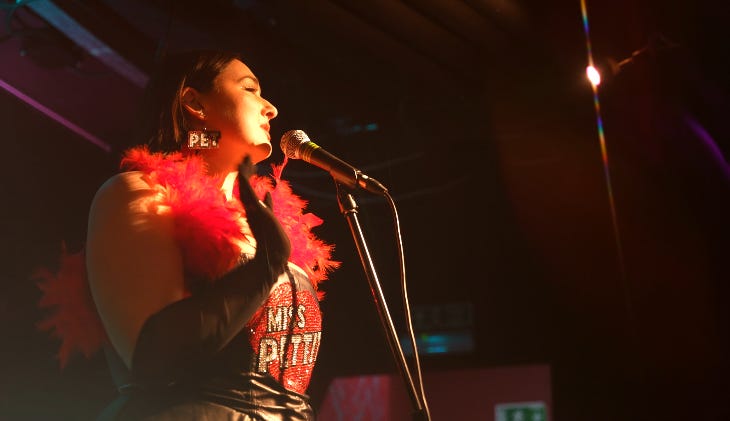 A performer on stage in front of a microphone wearing a red feather boa