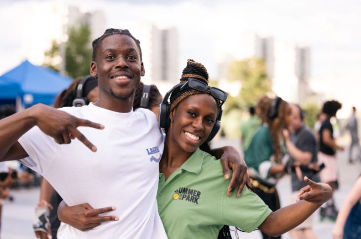 Two people, one wearing headphones, with their arms around each other smiling for the camera