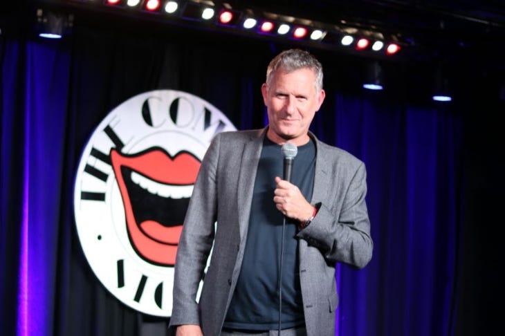 Adam Hills holding a microphone on stage at the Comedy Store