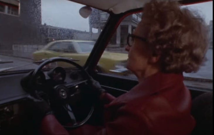 An elderly woman driving a car in a red leather jacket