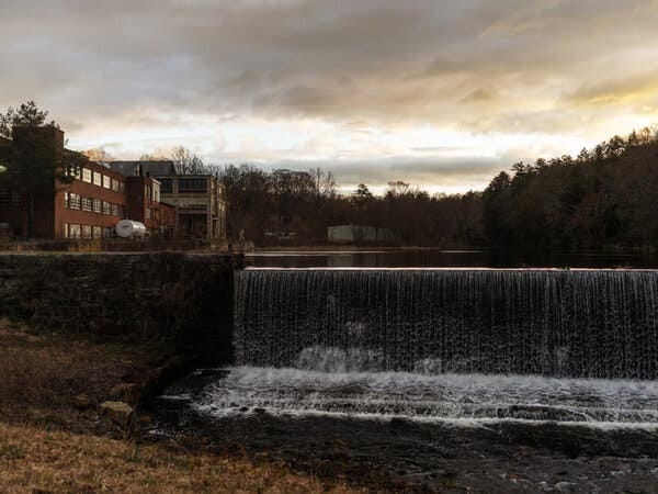 A brick building surrounded by woods and perpendicular to a small waterfall in the foreground.