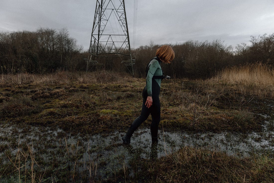 A photograph of a woman with a dark auburn bob walking through a peat bog, pictured side on. There is the tower of an eletricity pylon in the background.