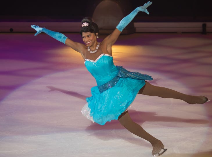 An ice skater dressed as Disney princess Tiana in a blue dress and gloves, and a diamond necklace