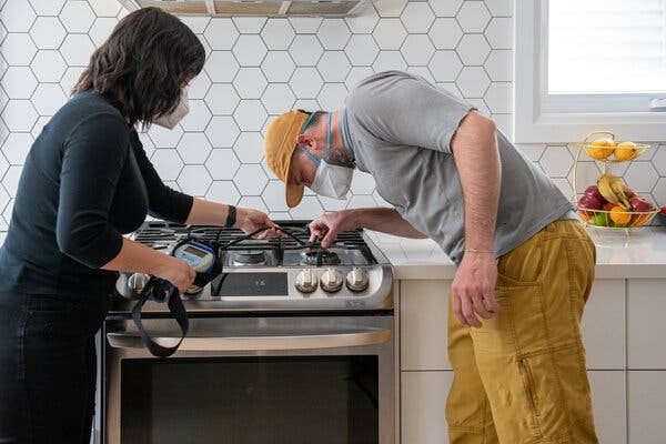 A female scientist in a black dress and a mask inserts a tube connected to a scientific instrument into the front burner of a gas stove, while a man in a yellow hat and yellow pants who is also wearing a mask peeks into the same burner.