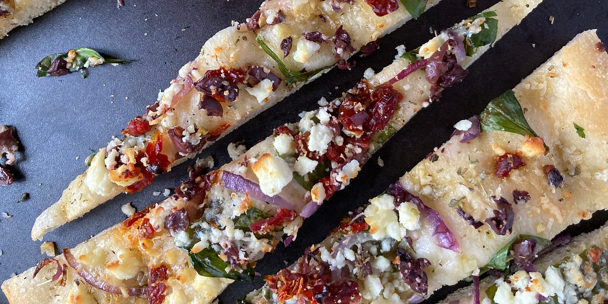 Focaccia bread topped with feta, sun-dried tomatoes, red onion and black olives on a black plate.