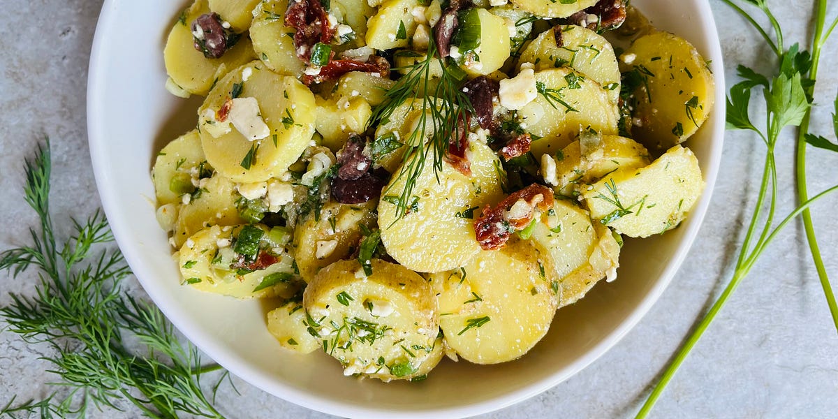 Potato salad with black olives, feta cheese, sun-dried tomatoes, dill and parsley in a white bowl.