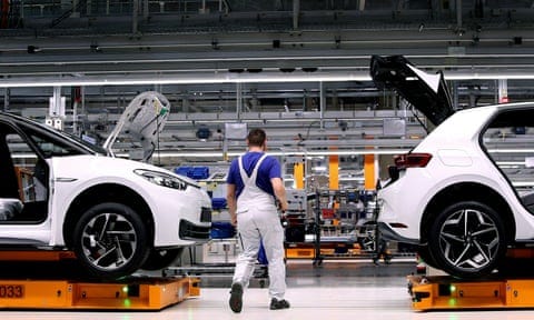 A German car worker in white dungarees attends to white coloured cars with their boots and bonnets open on an assembly line