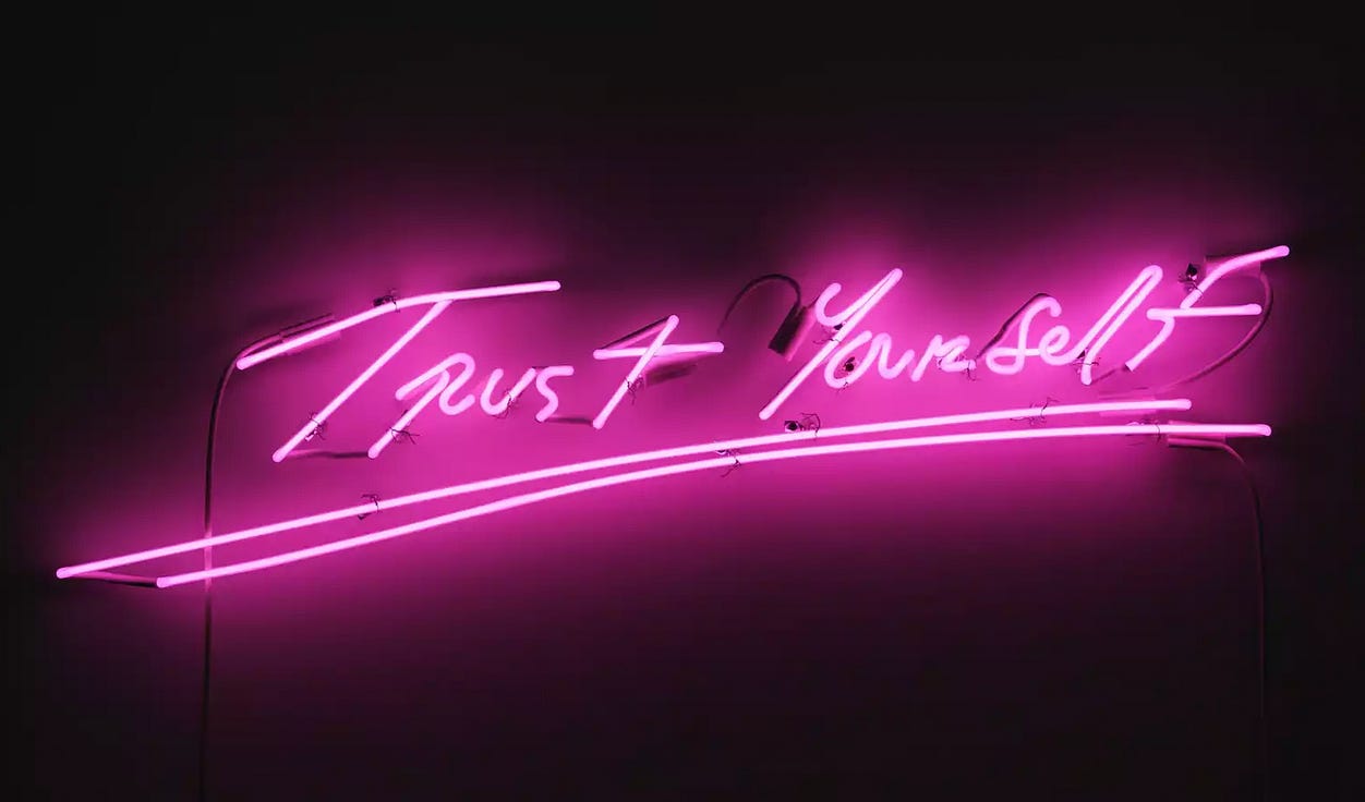 Black wall with a bright pink neon sign that says Trust Yourself in the artist's handwriting.