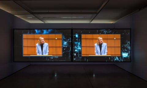 Two large screens hang side by side in a darkened gallery, each with the same image of Cate Blanchett at a lectern.