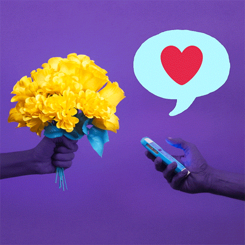 Video gif. A hand holds a phone and taps on it. A speech bubble rises up top that has a worried emoji, a blank face emoji, gritting teeth emoji, and a mad emoji in it. Other hand appears with a bouquet of yellow flowers. The hand types on the phone again and a speech bubble with a big heart pops up. 