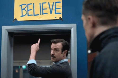 GIF from Ted Lasso: Ted Lasso points to the sign that reads BELIEVE.