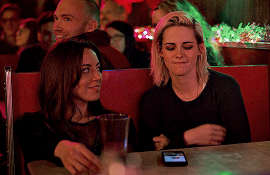 A gif of Aubrey Plaza and Kristen Stewart in the film Happiest Season. They are sitting next to each other in a booth at a bar. Aubrey smiles flirtatiously at Kristen who checks her phone. Aubrey looks away.