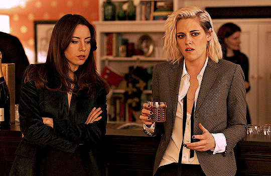 A gif of a scene in the film Happiest Season. Kristen Stewart's character Abby passes a drink to Aubrey Plaza's character Riley, who takes a sip.