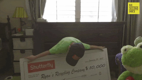 A young child with a neon green shirt and dark-colored hat holds up a giant check gifted by Shutterfly. It's worth $10,000 and made out to "Ryan's Recycling Company."
