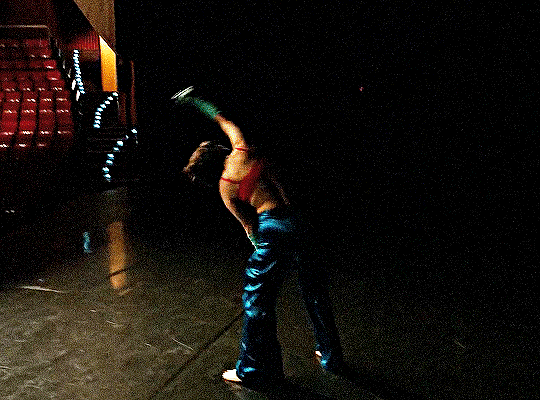 Mitski em Working For The Knife sozinha num palco, sem plateia, pulando freneticamente. Ela está de sutiã vermelho, calça e luvas azuis e descalça.