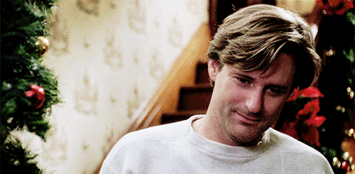 Bill Pullman in While You Were Sleeping standing on a staircase, wearing a white sweater, and smiling.