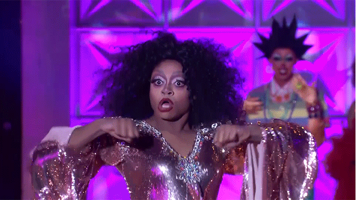 A gif of Heidi N Closet lipsynching for her life on RuPaul's Drag Race. She is on stage with another contestant and they are both dancing. She wears sparkly 70s style jumpsuit. She is shown doing a death drop - flinging herself onto the ground backwards.