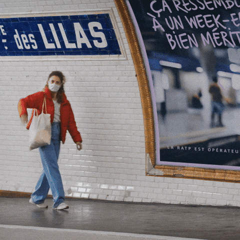 Una ragazza su un marciapede della metro parigina si sbraccia e saluta un ragazzo dall'altro lato del binario: lui sembra stranito e ricambia il saluto alzando dubbiosamente la mano.