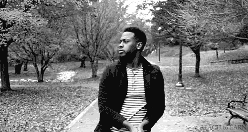 Black man with beard, tosses a bunch of fall leaves up in the air over his head using both hands energetically. He is standing on a path in a park. Black and white film