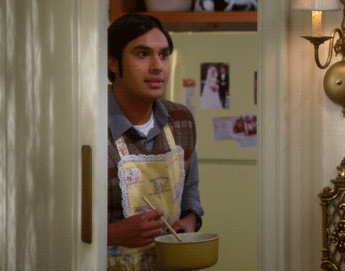Raj della serie The Big Bang Theory, ha in mano un pentolino e sorride leggermente mentre davanti a lui si chiude la porta della cucina.