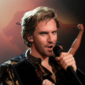 A gif of Dan Stevens as Alexander Lemtov. He is singing into a microphone while looking at the camera and winking.