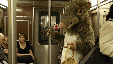 A human being in a large rat suit holding a wicker basket on the NYC subway. A few passengers look at the human-rat, who is holding a subway pole.