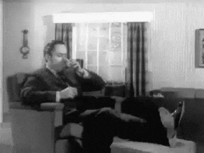 Black and white gif of a man sitting in a lounge at home who is enveloped by a giant suitcase and whisked away to a holiday destination.