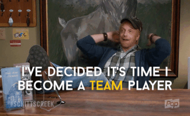 Roland from “Schitt’s Creek”, feet up on the desk, says, “I’ve decided it’s time I become a team player.”