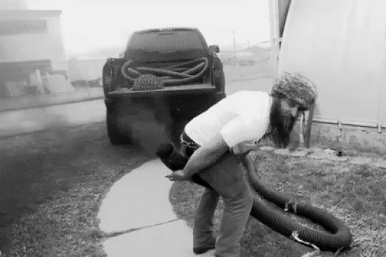 gif of a grinning bearded guy who has attached a long 5-inch diameter hose to the exhaust of a diesel pickup, and is holding the end of the hose between his legs so it appears the thick black diesel exhaust is coming from his butt