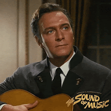 Christopher Plummer as Captain Von Trap in the Sound of Music. He's sitting with a guitar and he smiles and giggles a little.