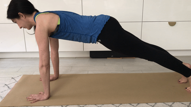 A woman with brown hair and a blue tank top performs a serratus push-up in plank position.