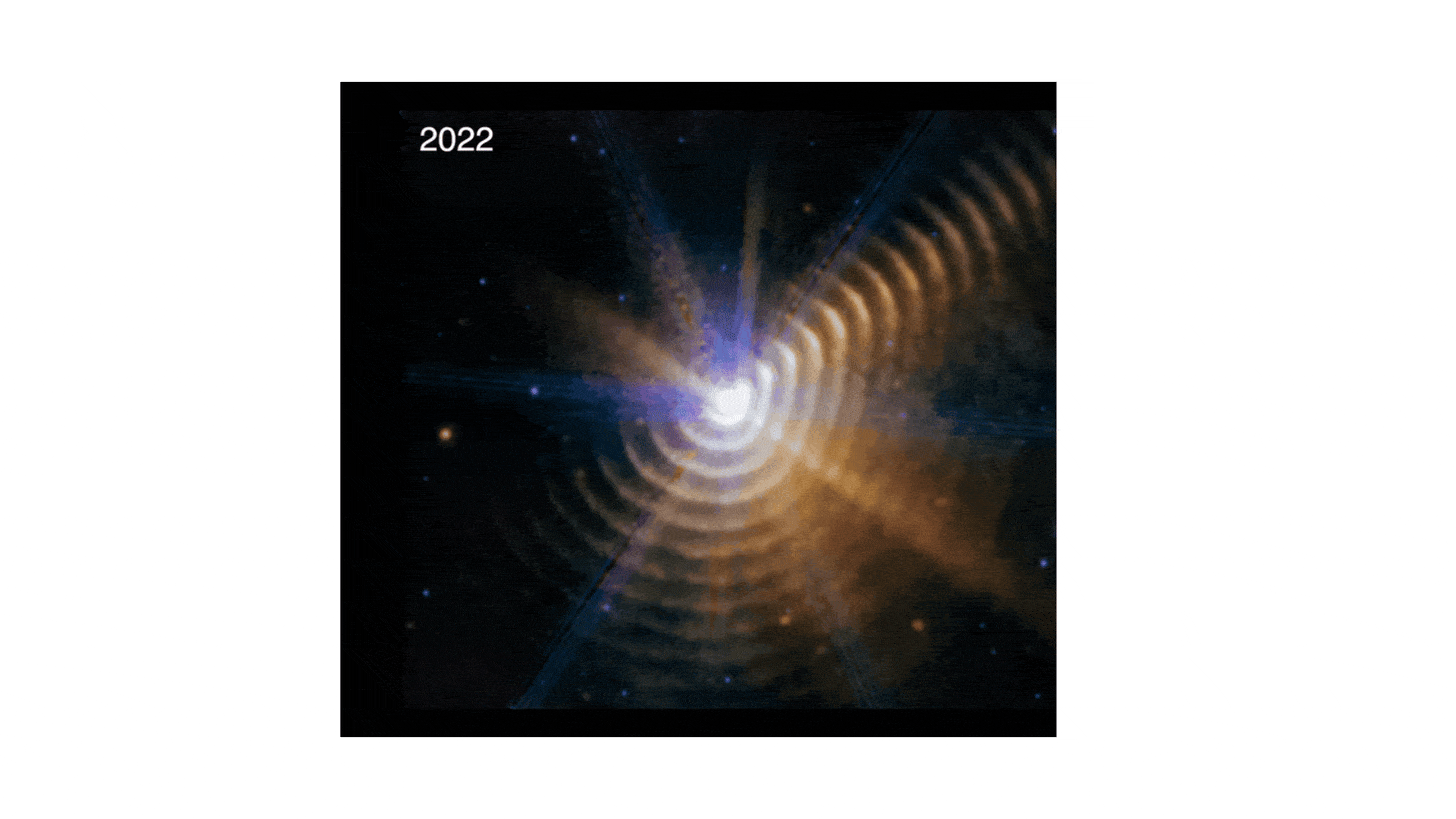 The video alternates between two James Webb Space Telescope images of the two-star system Wolf-Rayet 140, the first taken in 2022 and the second in 2023. Both show a bright white point of light surrounded by 17 regularly spaced, hazy dust shells at the bottom, right, and upper right. There is noticeably less color in the upper left. The central point, where the two stars are located, has a rough hexagon shape. By alternating between them, it’s clear that the dust shells are moving outward, becoming wider. The video alternates between two James Webb Space Telescope images of the two-star system Wolf-Rayet 140, the first taken in 2022 and the second in 2023. Both show a bright white point of light surrounded by 17 regularly spaced, hazy dust shells at the bottom, right, and upper right. There is noticeably less color in the upper left. The central point, where the two stars are located, has a rough hexagon shape. By alternating between them, it’s clear that the dust shells are moving outward, becoming wider.
