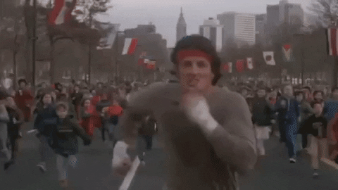 Movie gif. Sylvester Stallone as Rocky Balboa in Rocky runs down the middle of the road in a full sprint. A crowd of hundreds of people run behind him.