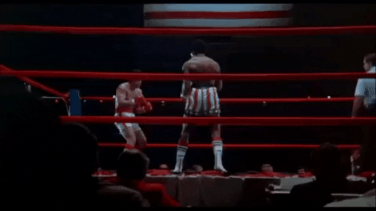 Due pugili sul ring circondato da corde rosse, uno con pantaloncini a stelle e strisce e l’altro con guantoni rossi, mentre si scambiano colpi in un match di boxe. Fotogramma tratto dal film Rocky.