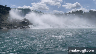 Niagara Falls, as seen from a boat approaching the water fall. The sky is blue, the sun is shining, and the spray from the water fall is splashing up into the air.