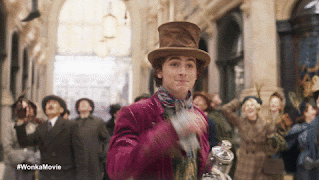 Timothée Chalamet as Wonka, is tipping his hat to the camera, whilst standing in a busy street with people walking by him.