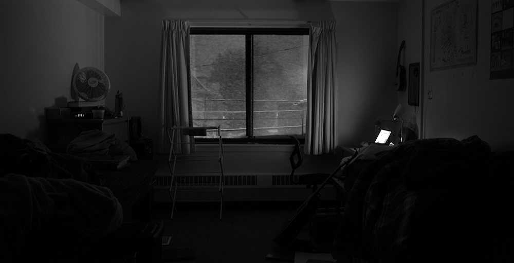 Dimly lit dorm room at night with a single window showing a quiet outdoor scene. A desk lamp illuminates a laptop screen, casting a glow on nearby objects. The room contains an unmade bed, scattered belongings, a fan, and a guitar hanging on the wall, creating a moody, introspective atmosphere.