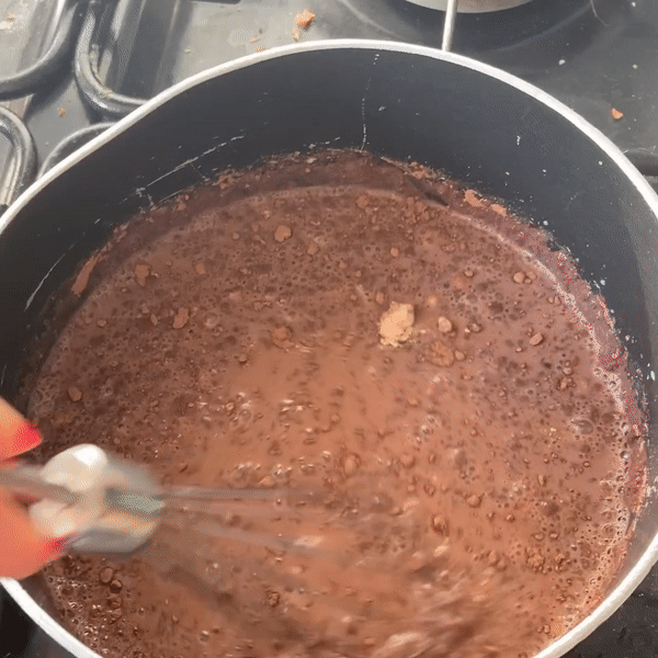 A moving gif showing a sauce pan on a hob. It is filled with hot chocolate, with not all the powder stirred in, being whisked. When the hand comes into view, it is a white hand with chipped red nail polish.