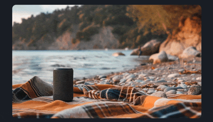 A speaker set on top of a blanket laid out on a the beach. There is w a shoreline in the background.