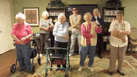 Video gif. A bunch of senior women stand in a living room, along with one young man in sunglasses and a tank top. A few of the older women have walkers, and they all stand together doing the macarena.