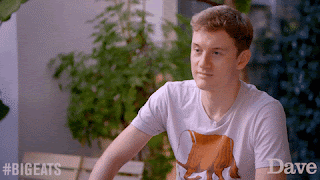 James Acaster, looks at the camera, pulls a face that shows a picture of confusion and amusement, and then looks away. He's wearing a white t shirt with what looks like an illustrated loaf of bread with legs on it.